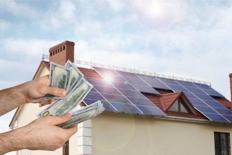 A person holding money in front of a house with solar panels on the roof.